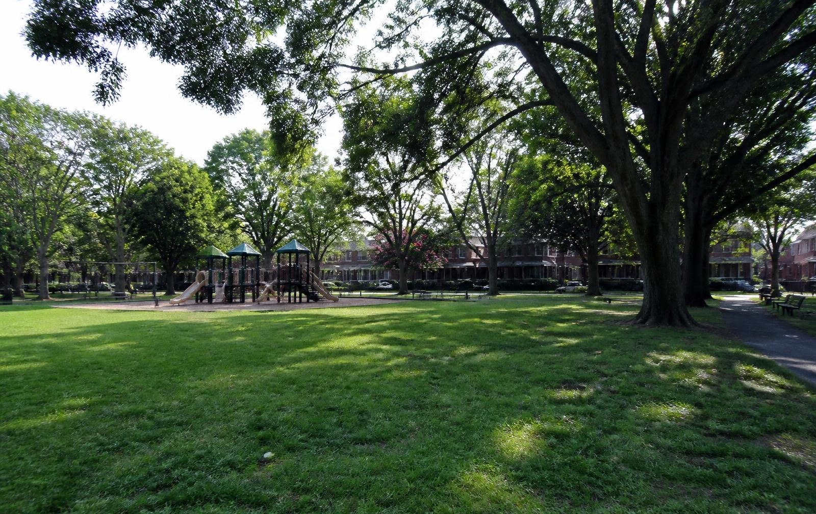 Open shaded lawn and small playground at a neighborhood pocket park in Charlotte, framed by mature willow oaks with brick houses beyond the tree line