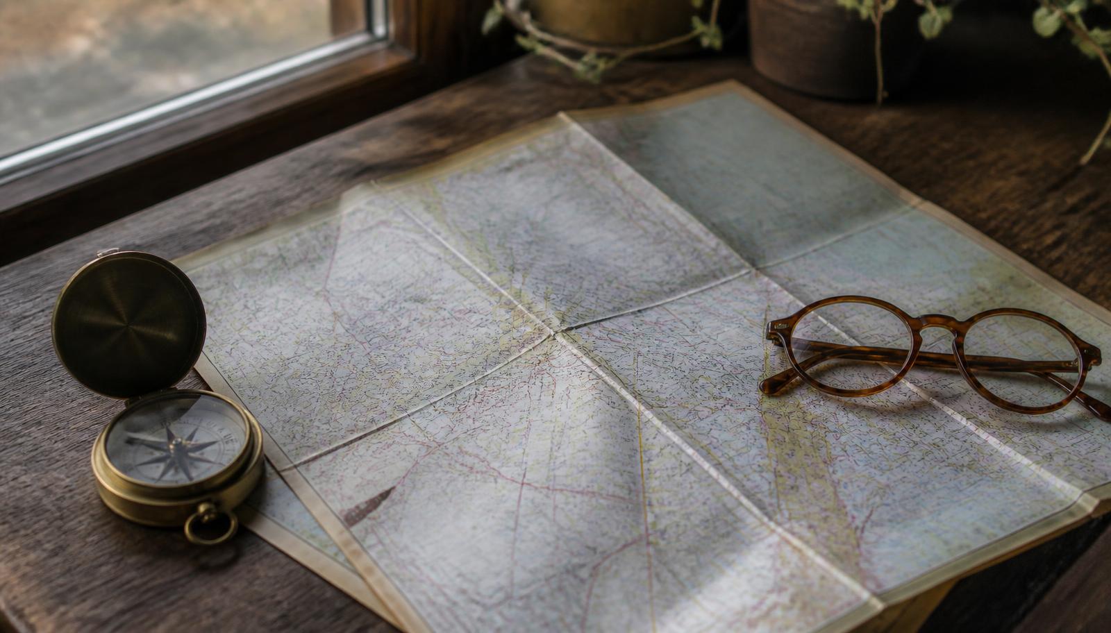 Folded city map, brass compass, and reading glasses on a wooden table
